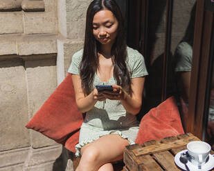 Attractive Asian female surfing cellphone while sitting at table with empty cup of coffee on terrace of cafeteria in summer - ADSF35920