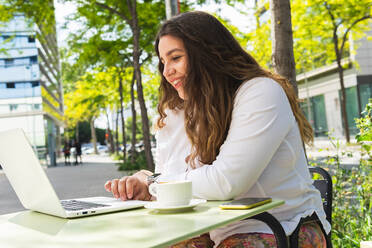 Seitenansicht einer zufriedenen Frau, die auf ihrem Netbook surft, während sie mit einem Kaffee auf der Terrasse einer Cafeteria im Freien sitzt - ADSF35921
