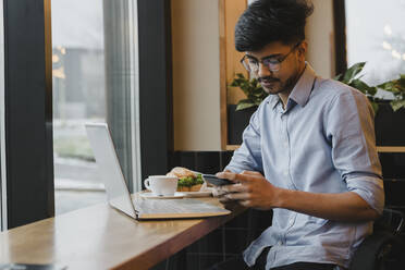 Junger Freiberufler mit Smartphone und Laptop in einem Cafe - OSF00322