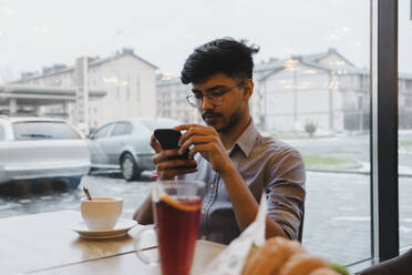 Junger Mann mit Smartphone am Fenster eines Cafés - OSF00345