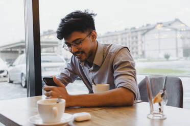 Happy man using smart phone by window in cafe - OSF00346