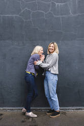 Young woman laughing with mother by wall on footpath - ASGF02526