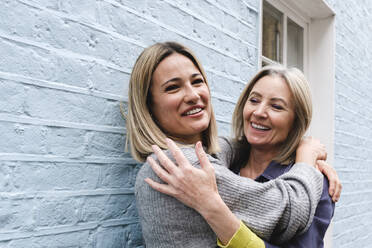 Happy young woman embracing mother in front of blue wall - ASGF02532