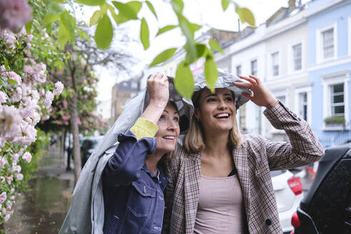 Smiling mother and daughter under windbreaker in rain weather - ASGF02565