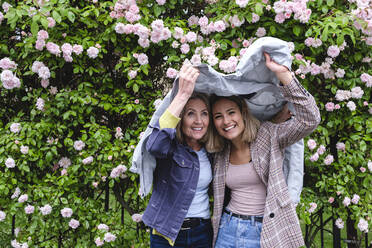 Smiling mother and daughter under windbreaker in front of flowers - ASGF02566