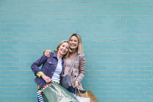 Happy mother and daughter with shopping bags in front of blue wall - ASGF02574