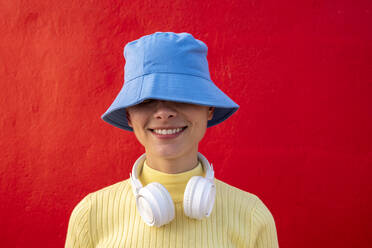 Smiling woman with headphones and blue bucket hat in front of red wall - VPIF06659