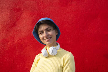 Smiling woman with headphones and bucket hat in front of red wall - VPIF06661