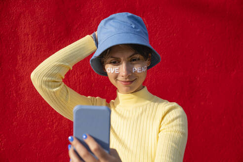 Frau mit Eimer Hut nimmt Selfie auf Smartphone vor roter Wand - VPIF06662