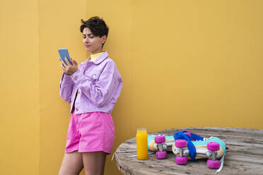 Beautiful woman using mobile phone leaning on yellow wall by juice and roller skates on table - VPIF06674
