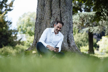 Lächelnder Geschäftsmann mit Mobiltelefon vor einem Baum - JOSEF11082