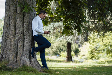 Älterer Geschäftsmann, der ein Smartphone benutzt und sich im Park an einen Baumstamm lehnt - JOSEF11087