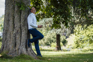 Mature businessman holding mobile phone leaning on tree trunk in park - JOSEF11088