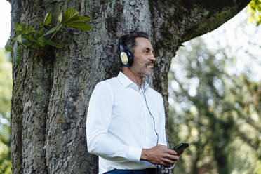 Geschäftsmann, der ein Mobiltelefon hält und über Kopfhörer Musik hört, lehnt sich an einen Baumstamm - JOSEF11089