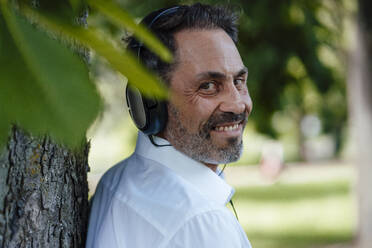 Lächelnder Geschäftsmann, der über Kopfhörer Musik hört und sich an einen Baumstamm lehnt - JOSEF11095