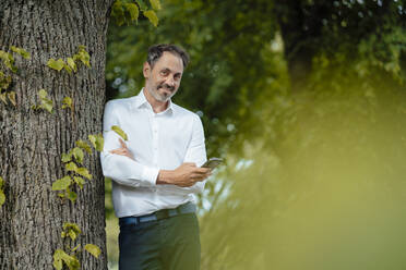 Smiling mature businessman holding smart phone standing by tree - JOSEF11097