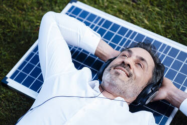 Smiling businessman with hands behind head lying on solar panel listening music through headphones - JOSEF11098