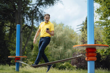 Surprised man looking at smart phone balancing on tightrope in park - JOSEF11110