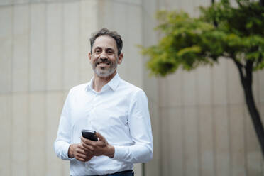 Smiling businessman with smart phone in front of building - JOSEF11115