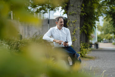 Älterer Geschäftsmann mit Smartphone sitzt auf einer Bank im Park - JOSEF11126