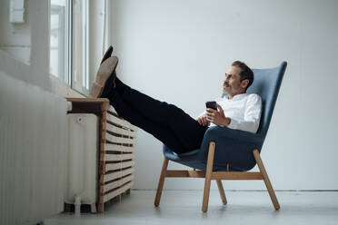 Geschäftsmann mit Smartphone auf einem Sessel sitzend und durch ein Fenster im Büro blickend - JOSEF11136