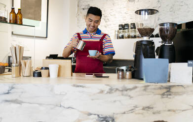 Lächelnder junger Mann mit Kaffeebecher und Tasse bei der Arbeit in einem Cafe - JCCMF06684