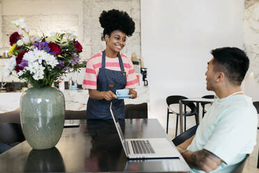 Eine lächelnde Kellnerin serviert einem Kunden, der mit einem Laptop in einem Café sitzt, Kaffee - JCCMF06707