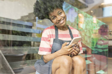 Glückliche junge Frau mit Smartphone in einem Café sitzend, gesehen durch ein Glasfenster - JCCMF06715