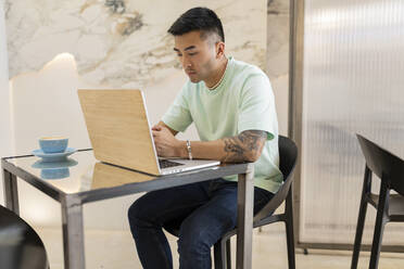 Junger Mann sitzt mit Laptop am Tisch in einem Café - JCCMF06719