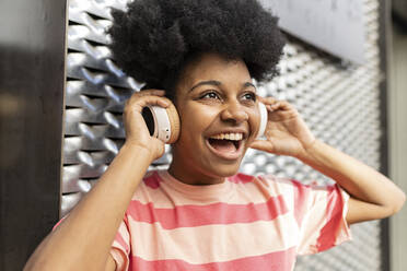 Fröhliche junge Frau, die vor einer Wand Musik über drahtlose Kopfhörer hört - JCCMF06760