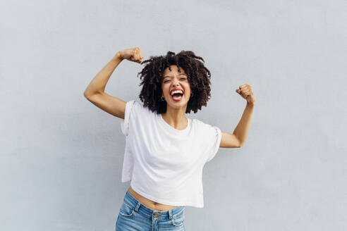 Cheerful young woman flexing muscles in front of white wall - MEUF06927