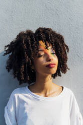 Junge Frau mit geschlossenen Augen genießt Regenbogen Sonnenstrahl vor der Wand - MEUF06928