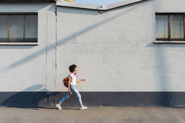 Junge Frau mit Rucksack, die auf einem Fußweg an einer Mauer vorbeiläuft - MEUF06933
