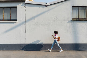 Junge Frau, die ein Smartphone benutzt und an einer Mauer auf dem Gehweg entlangläuft - MEUF06934