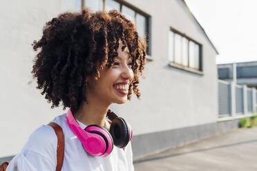 Happy young woman with curly hair wearing wireless headphones on sunny day - MEUF06936