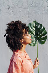 Young woman with eyes closed holding monstera leaf in front of wall - MEUF06943