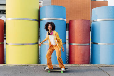 Happy woman standing on skateboard in front of colorful pipes - MEUF06974