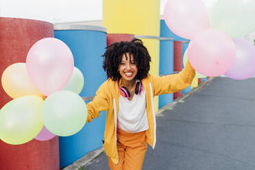 Smiling woman holding colorful balloons in front of pipes - MEUF06988