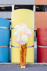 Junge Frau bedeckt ihr Gesicht mit bunten Luftballons vor einem Rohr - MEUF06990