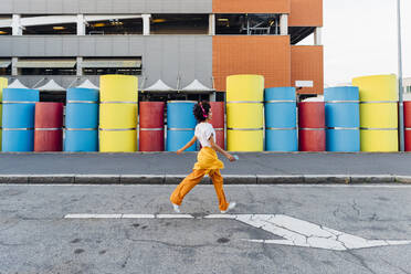 Woman listening music through wireless headphones walking on road by multi colored pipes - MEUF07007