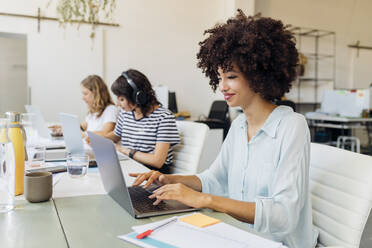 Smiling businesswoman with curly hair working on laptop in office - MEUF07068