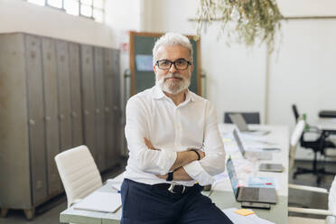 Confident businessman with arms crossed sitting on table in office - MEUF07087