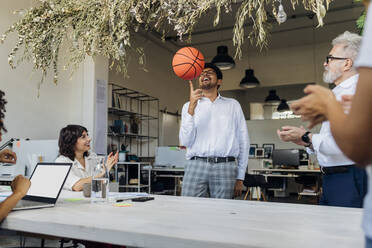 Playful businessman spinning basketball on finger by colleagues cheering in office - MEUF07157