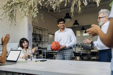 Colleagues congratulating businessman holding basketball at office - MEUF07158