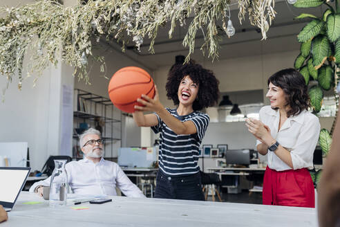 Glückliche Geschäftsfrau hält Basketball von Kollegen im Büro - MEUF07160