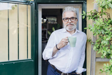 Älterer Geschäftsmann mit Kaffeetasse an der Tür lehnend - MEUF07169