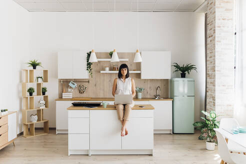 Woman sitting with laptop on kitchen island at home - MEUF07193