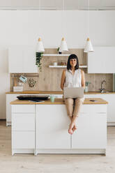 Woman with laptop sitting on kitchen island at home - MEUF07194