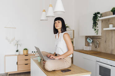 Thoughtful freelancer sitting on kitchen island with laptop at home - MEUF07197