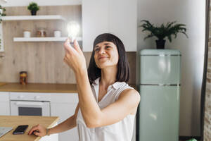 Smiling woman holding illuminated light bulb in kitchen at home - MEUF07203